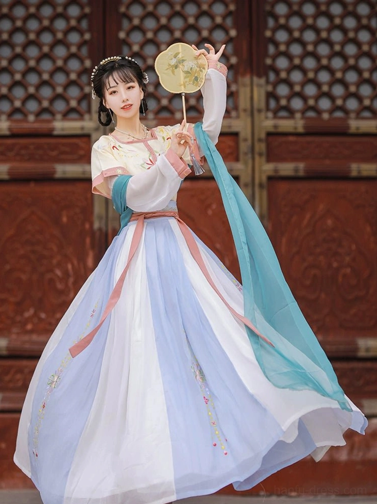 Three-quarter view of a cream yellow and pink Tang Dynasty Hanfu Qiyao Ruqun with floral embroidery, flowing cyan sash, and round silk palace fan beside a carved red wooden door