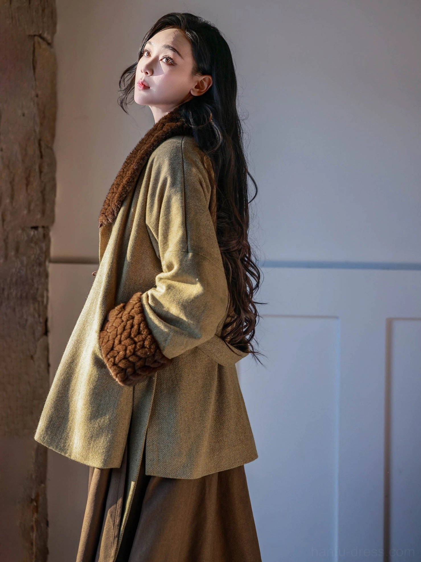 Side view of a model wearing an olive green Tang Dynasty inspired wool jacket with dark brown fuzzy trim and a brown pleated wrap skirt