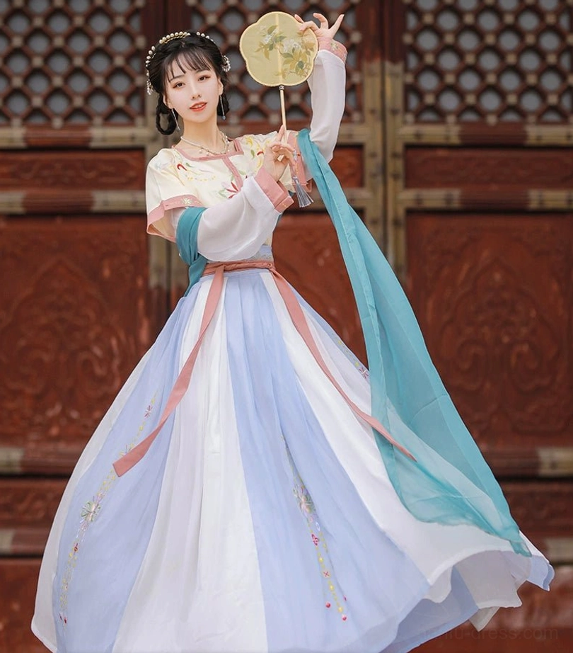 Low three-quarter view of a cream yellow Tang Dynasty style Hanfu set with pink trim, embroidered Qiyao Ruqun skirt, cyan sash, and round silk fan