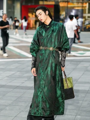 Full-body side view of a male model wearing a deep forest green Tang Dynasty Hanfu robe with flowing long silhouette, jacquard satin fabric, embroidered cuffs, ornate metal waist belt, and modern handbag on an urban street