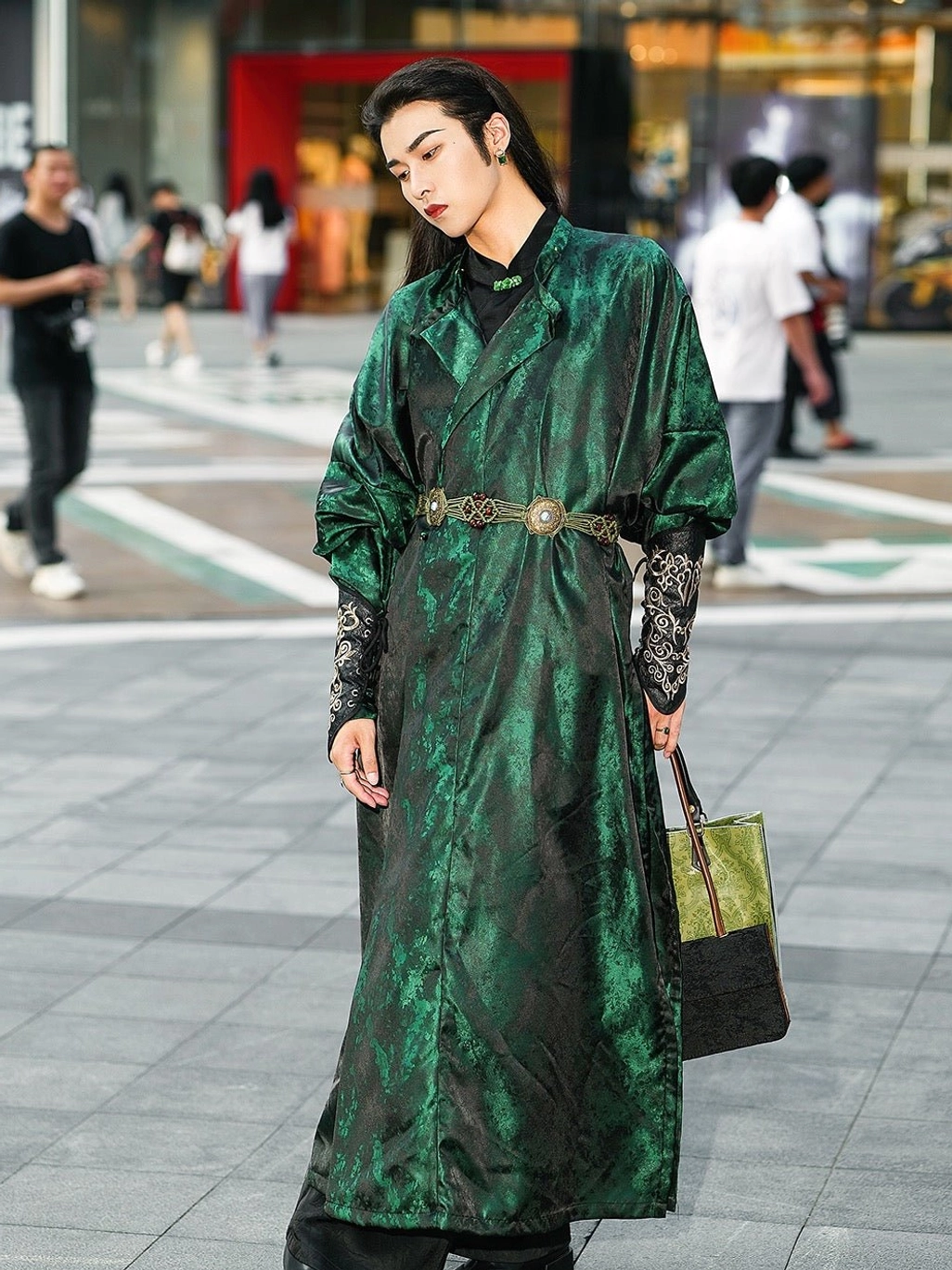 Full-body side view of a male model wearing a deep forest green Tang Dynasty Hanfu robe with flowing long silhouette, jacquard satin fabric, embroidered cuffs, ornate metal waist belt, and modern handbag on an urban street