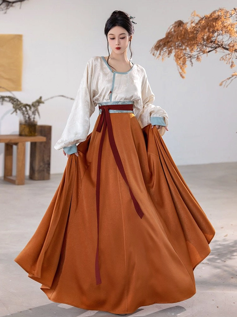 Three quarter front view of Tang Dynasty modern Hanfu set showing ivory jacquard top, light blue trim, and soft flowing burnt orange satin skirt