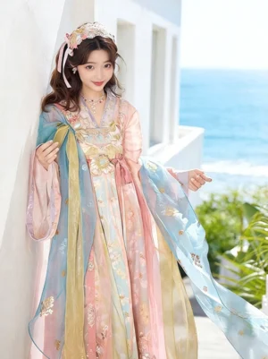 Front three-quarter view of a pastel Tang Dynasty Qixiong Ruqun Hanfu dress with gradient colors, floral embroidery, wide sleeves, and flowing golden scarves worn by a woman outdoors