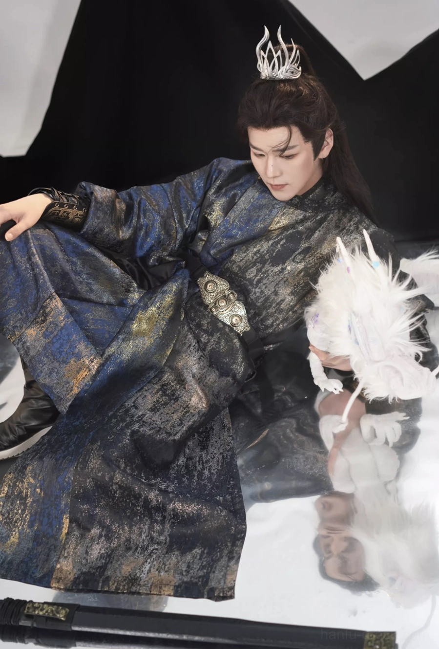 Full sitting view of a Tang Dynasty inspired warrior hanfu for men with detailed dark jacquard textile, long black outer vest, navy robe, decorative belt, and mountain fantasy backdrop