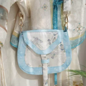 Front overhead view of a Tang-inspired blue floral and rabbit embroidered Chinese crossbody saddle bag styled with beige Hanfu, sky-blue fabric edging, and gold-tone hardware