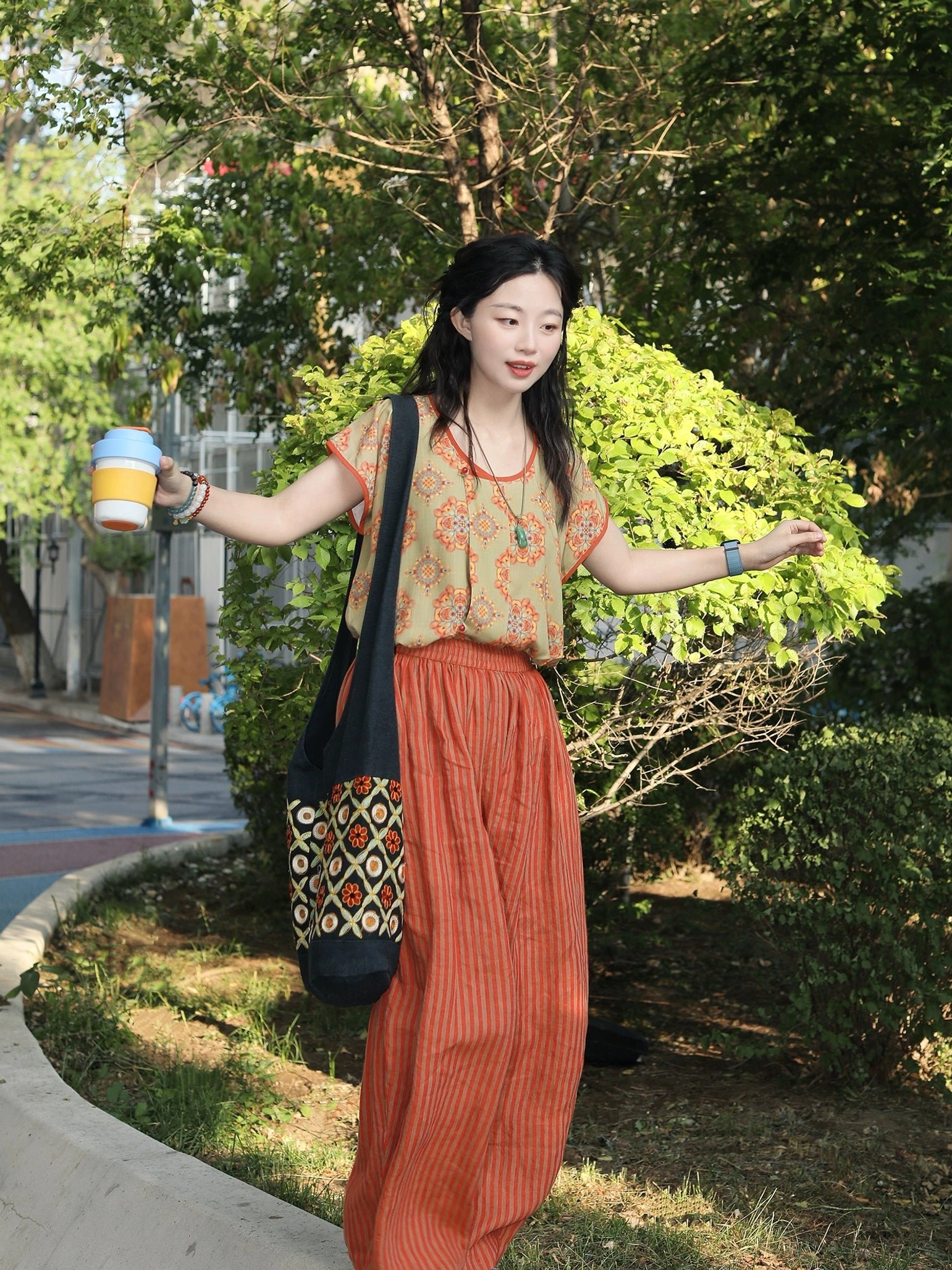 Three-quarter side view of a woman in a Tang-inspired Hanfu half-sleeve top and high-waisted rust-red striped linen pants carrying a matching patterned canvas tote