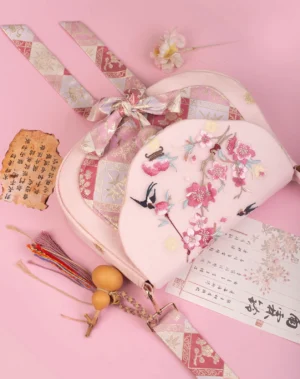Angled view of blush pink embroidered Chinese bag with flower bird flap embroidery, gold geometric jacquard patchwork, bow shoulder strap, and tassel charm