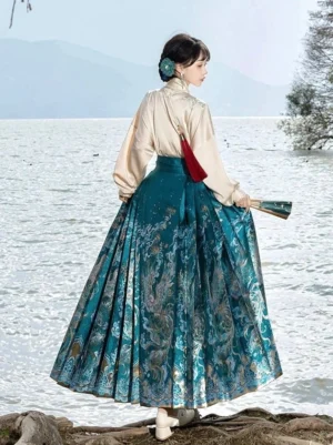 Back view of a teal dragon jacquard mamianqun Hanfu outfit with light cross-collar blouse and red waist pouch accessory beside a lake