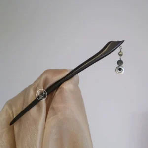 Handheld view of a natural wood Chinese hair stick with shell tassel displayed beside light apricot Hanfu fabric