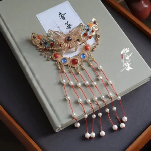 Traditional Chinese butterfly tassel back hair pin with gold filigree frame red tassels white pearl pendants and colorful gemstone details displayed on a grey book