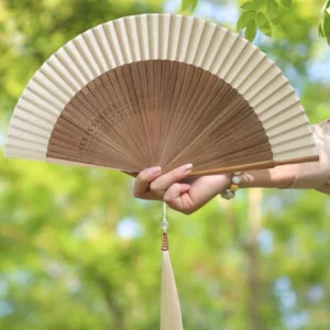 Traditional Chinese folding hand fan with carved bamboo ribs, pleated off-white fan surface, two-tone design, and decorative tassel held outdoors against green foliage