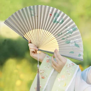 Traditional Chinese folding fan with grey floral and leaf artwork, smooth yellow bamboo ribs, decorative tassel, and jade-style accent held with embroidered Hanfu in an outdoor setting