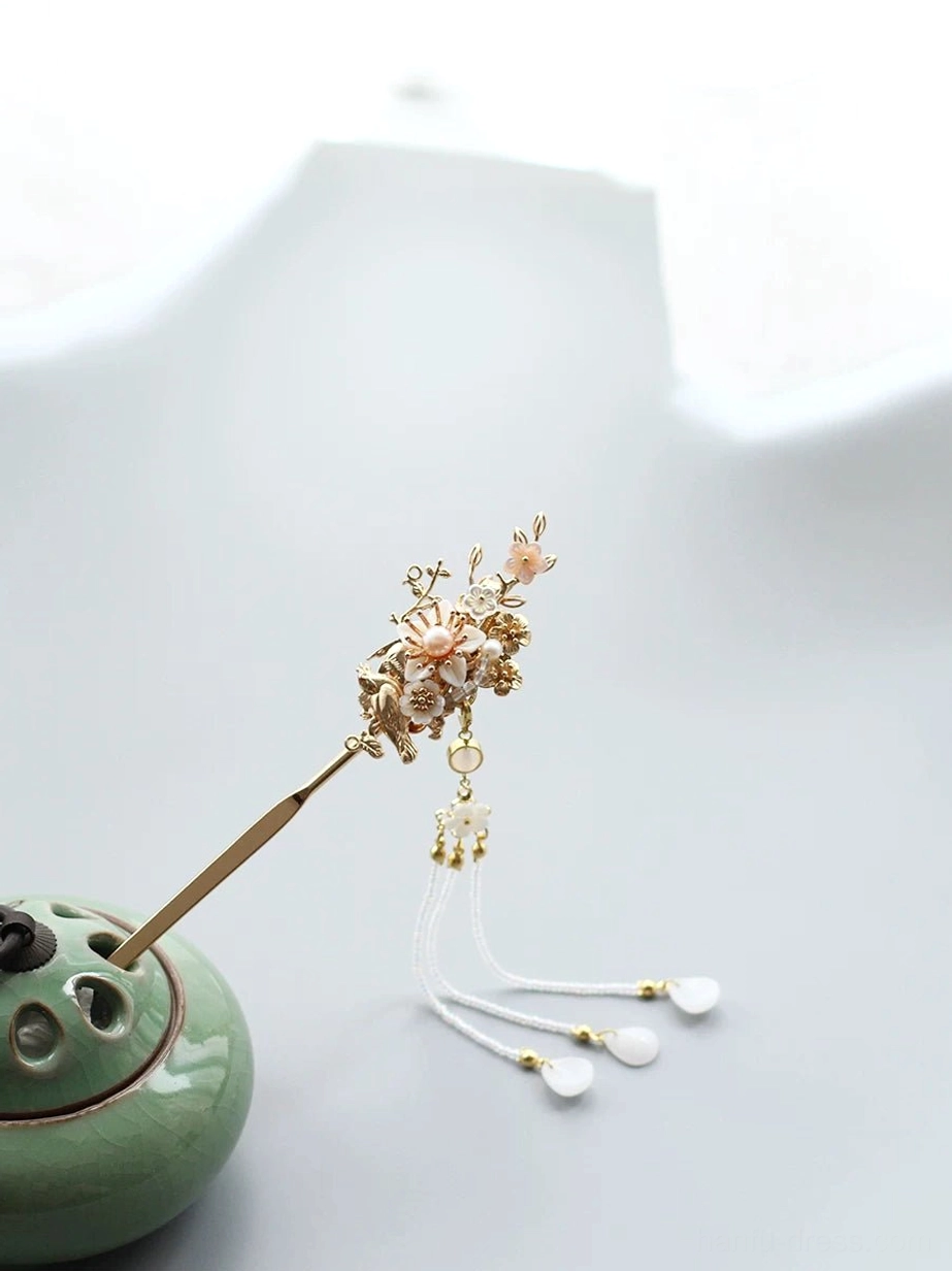 Traditional Chinese hair accessory with gold floral ornaments, pink flower accents, white tassel, and gold-capped pendants beside a green incense burner