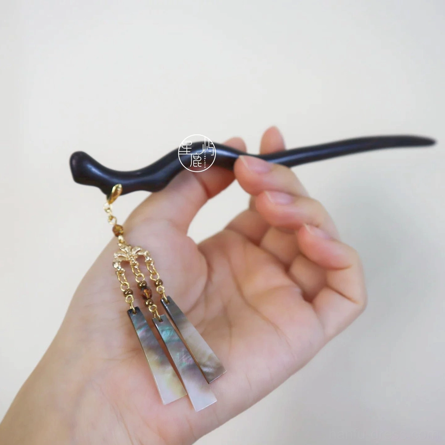 Hand holding a traditional Chinese hair stick with a wavy dark wooden body, detailed beaded tassel, and decorative shell pendant