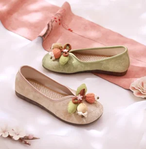 Pair of traditional Chinese Hanfu shoes in beige and sage green displayed on white satin with a pink Hanfu sash and peach blossom decorations