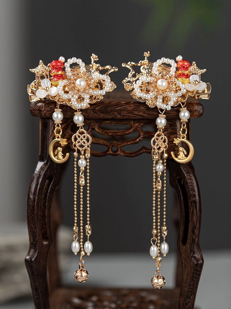 Pair of traditional Chinese Hanfu tassel hairpins with gold floral details faux pearl clusters red flower accents and long chain moon charms displayed on a carved wooden stool