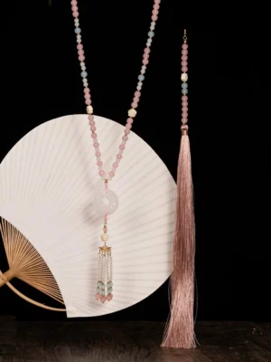 Traditional Chinese long tassel necklace displayed on a wooden table with a white folding fan, pastel beaded chain, white jade-style donut pendant, metal tassel charm, and matching pink tassel accessory