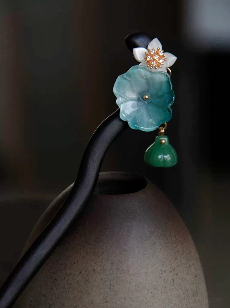 Traditional Chinese lotus hair stick leaning against a ceramic jar, featuring a black curved wooden shaft, translucent blue lotus, white flower, and green pendant