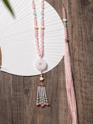 Close-up of a traditional Chinese necklace with pink beaded chain, white jade-style donut pendant, filigree tassel charm, colorful beads, and long pink tassel beside a white folding fan