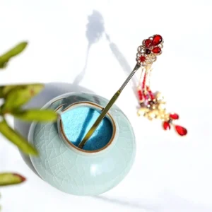 Traditional Chinese ruby red tassel hairpin displayed on celadon jar with gold tone pin and flowing beaded tassels