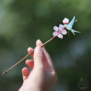Side view of a slender traditional Chinese hair stick with miniature peach blossoms and a small blue swallow decoration held in a hand outdoors
