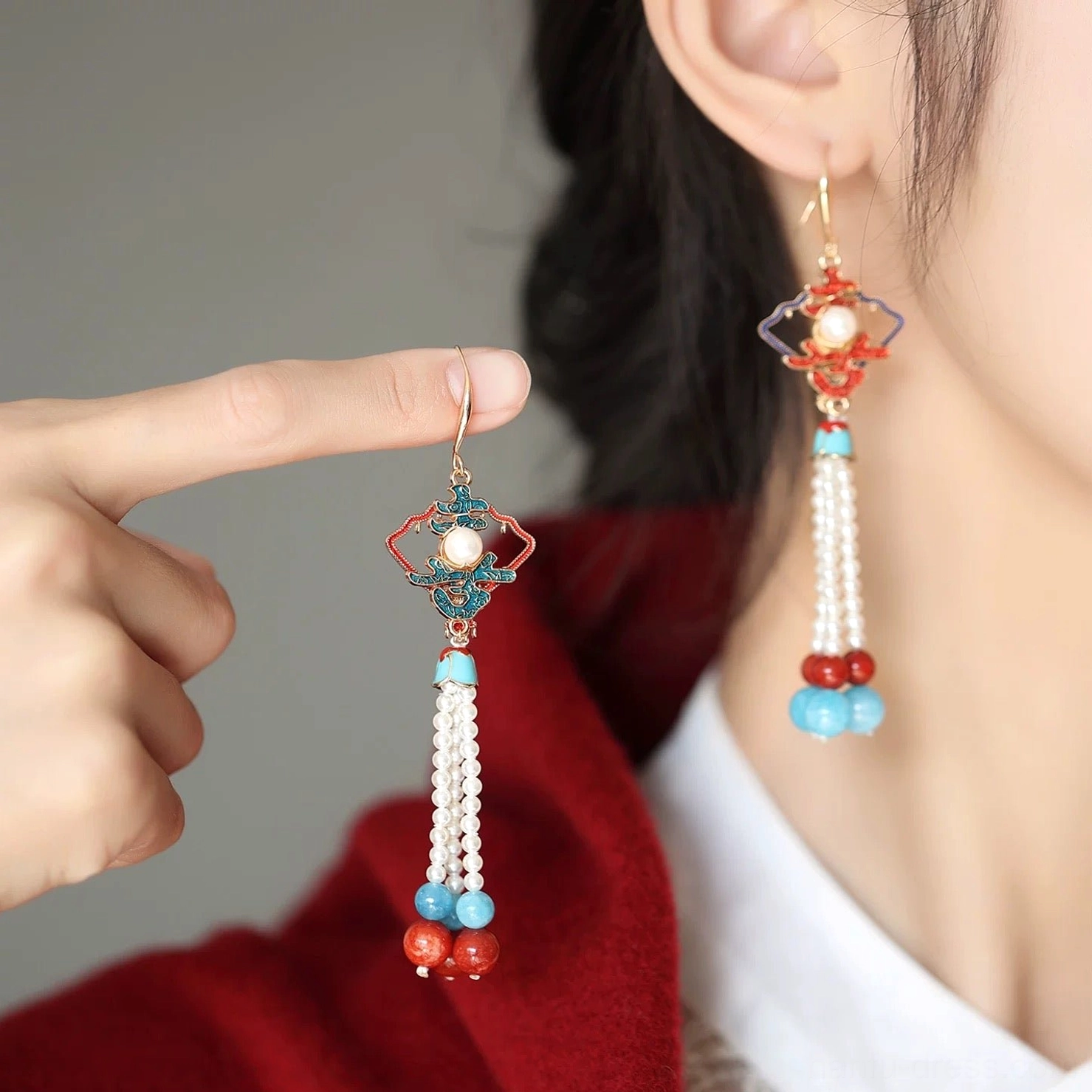 Close-up of a traditional Chinese tassel earring held by hand and worn on the ear, showing gold hook, enamel motif, pearl center, long pearl tassel, and layered bead pendants