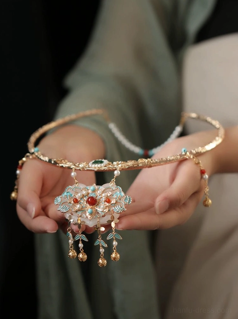 Close-up of traditional Chinese wedding necklace held in front of light green Hanfu clothing, featuring a gold frame, red center stone, enamel details, and bell tassels