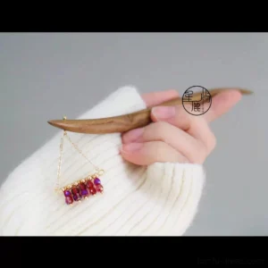 Close-up handheld view of a traditional Chinese wooden hair stick showing its smooth wood texture, gold-tone chain, and wine red crystal decoration