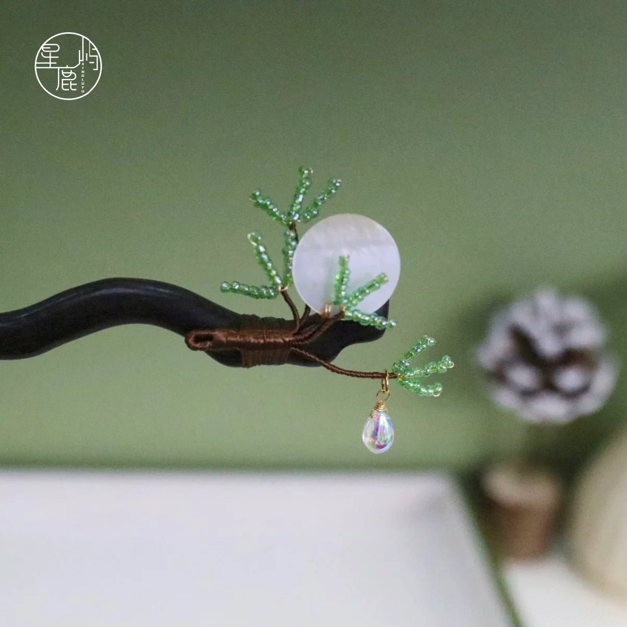Traditional Chinese wooden hair stick displayed against a green background with pine cones, showing the curved dark wood, green pine decoration, shell disc, copper wire, and iridescent pendant