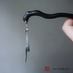 Side close-up of a traditional Chinese wooden hair stick held in hand, showing the irregular matte black wood texture, extended silver chain, green pendants, and decorative tassel