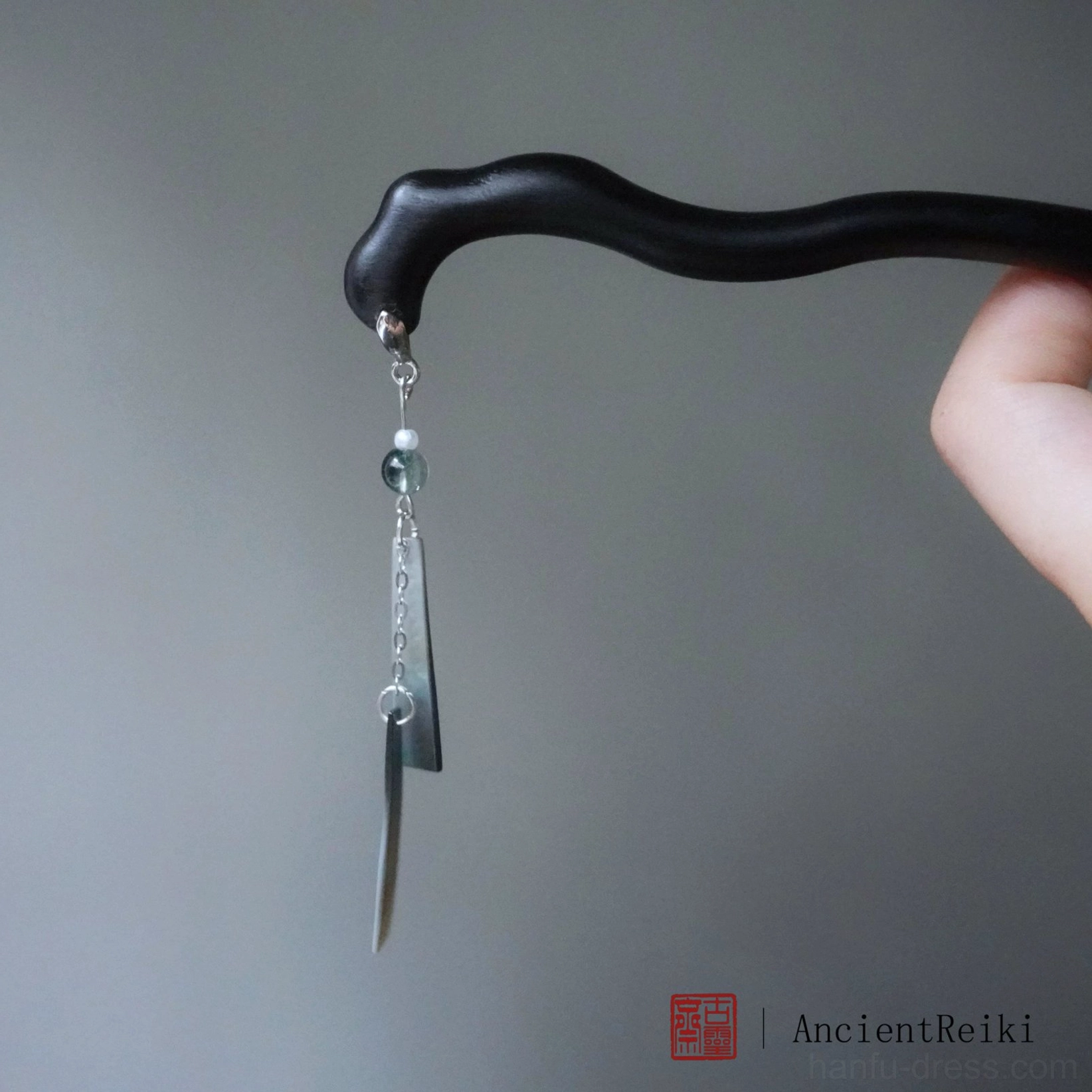 Side close-up of a traditional Chinese wooden hair stick held in hand, showing the irregular matte black wood texture, extended silver chain, green pendants, and decorative tassel