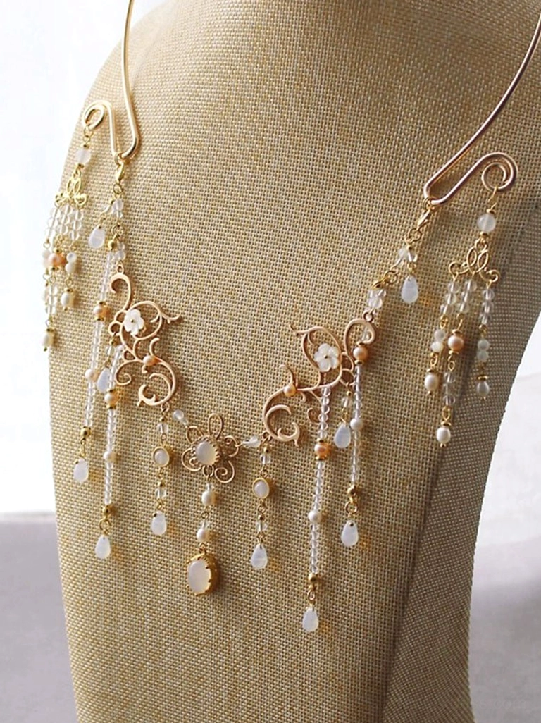 Traditional Chinese gold yingluo necklace on a beige mannequin featuring central floral filigree, layered dangling pendants, and a prominent white teardrop charm