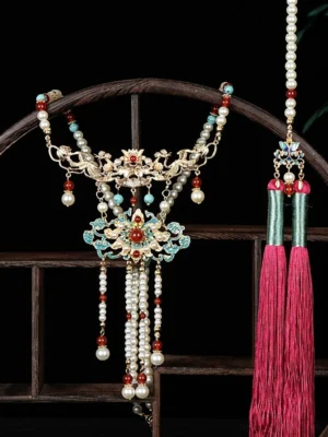Front view of a traditional Chinese crane-style hanfu necklace with gold-toned cloisonne floral decorations, pearl-style beads, red accents, and rose-red tassels on a vintage wooden display