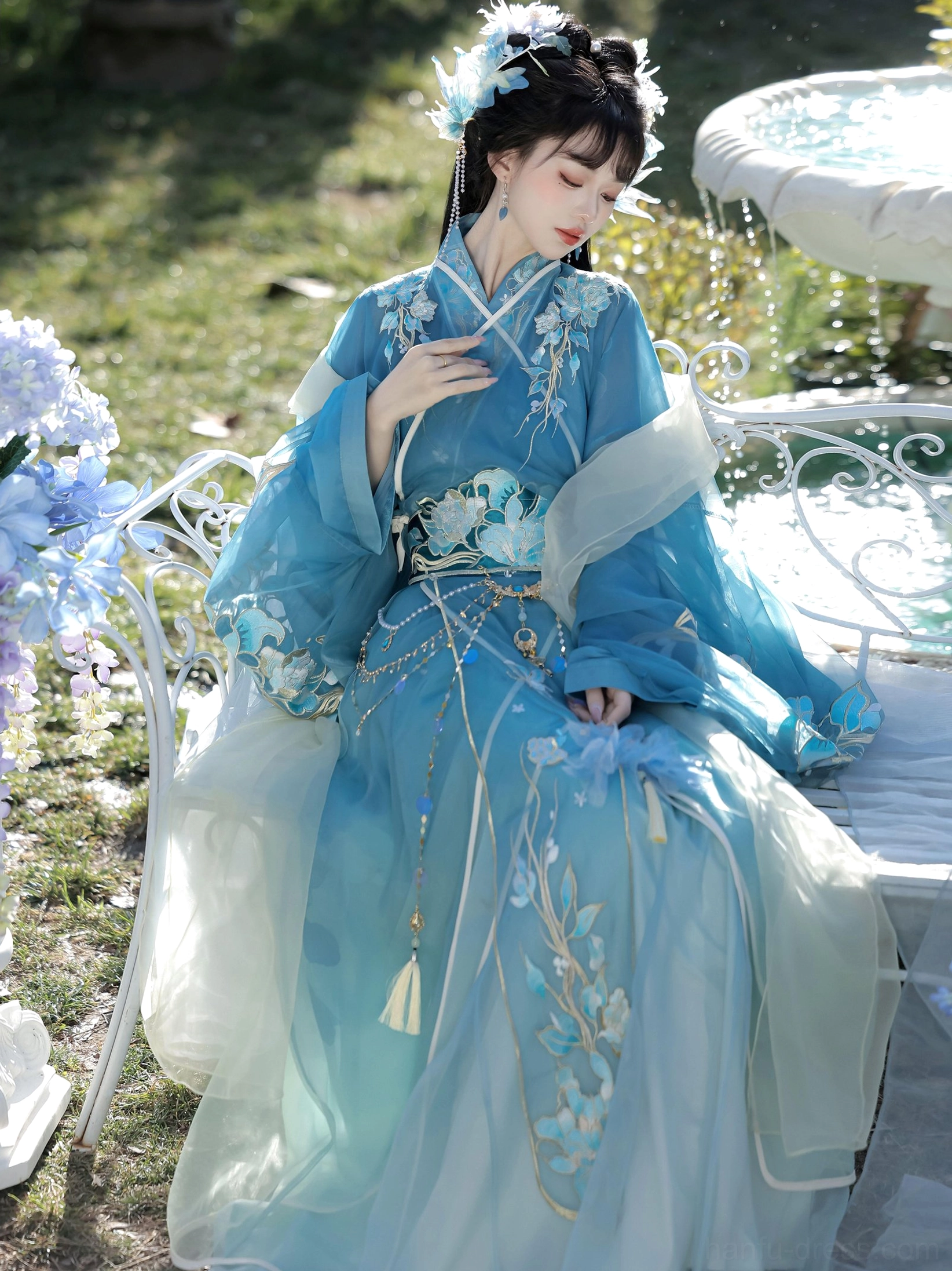 Traditional Hanfu robe shown seated in a garden with floral embroidery, layered light cyan sheer skirt, and flower-patterned decorative waist belt