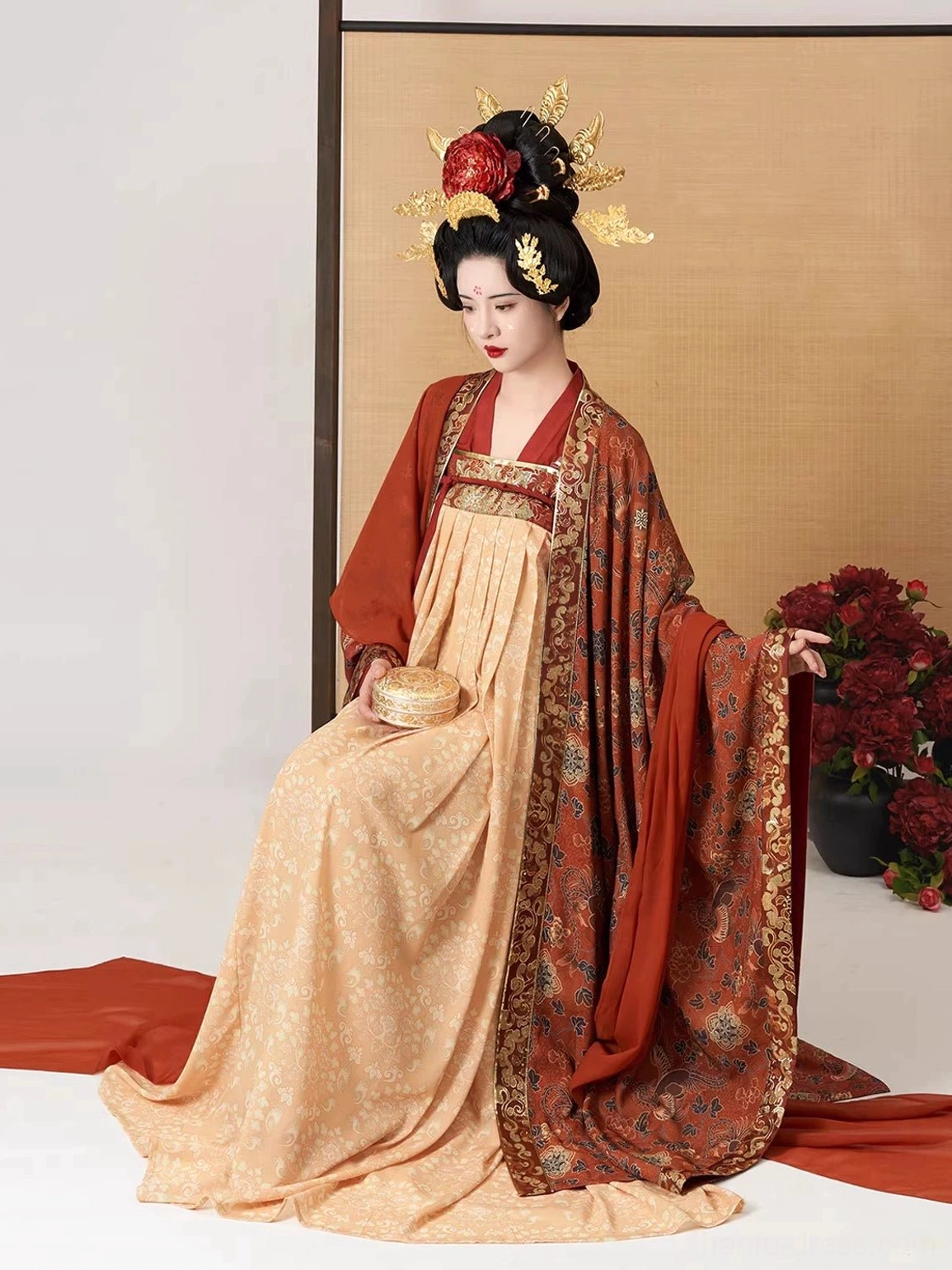 Seated side view of a model wearing a traditional Tang Dynasty Qixiong Ruqun Hanfu with a floor-length draped skirt, rust-red patterned outer robe, bamboo screen, and red peonies