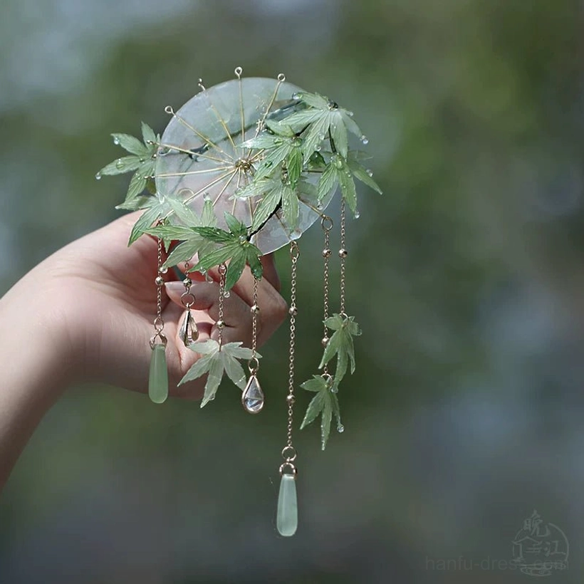 Front view of a Chinese maple leaf hair stick with transparent round pendant, gold chains, jade-style ornaments and crystal teardrops held in hand