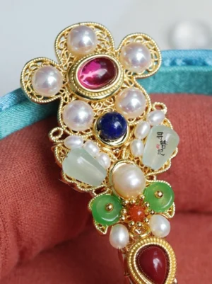 Close-up of Treasure Wind traditional Chinese hairpin featuring ornate gold-tone floral filigree, pink-red center stone, pearl-like beads, blue and green jade-style ornaments