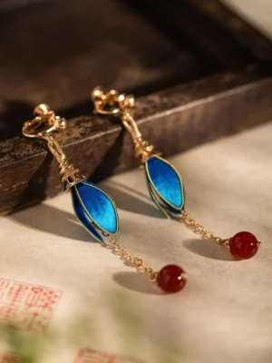 Blue flower bud clip-on Chinese earrings with glossy petal charms, red dangling bead pendants, and gold-tone fittings on a dark wooden tray