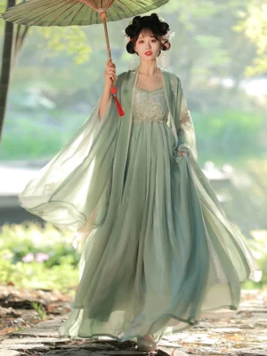 Front full-body view of a dusty green Tang Dynasty Hanfu Heziqun dress set with wide embroidered sleeves, flowing skirt, traditional hairstyle, and green oil-paper umbrella beside a lakeside woodland path