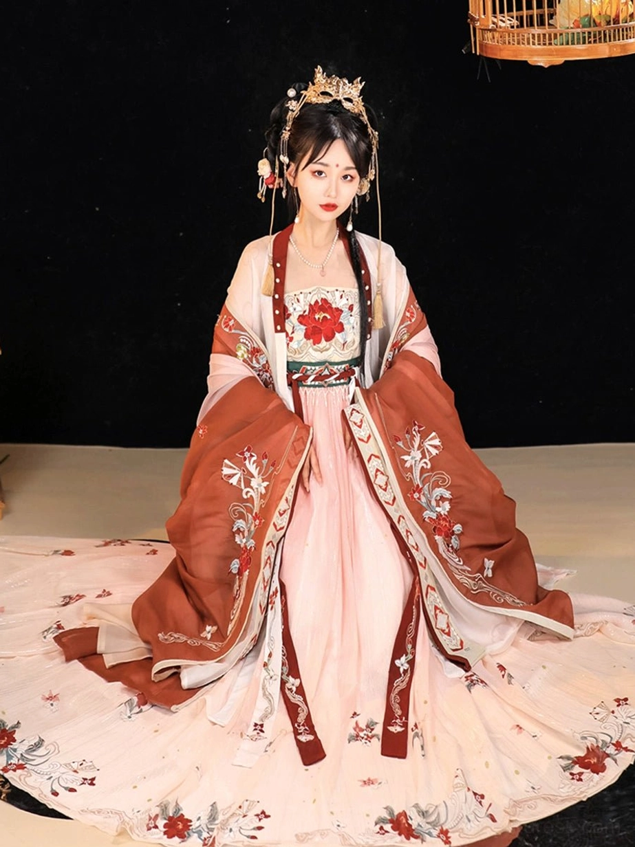 Seated model wearing the pink gradient Twilight Tang Dynasty Hanfu Heziqun dress with reddish-brown wide-sleeve coat, floral embroidery, coordinated chest panel, and golden hair crown