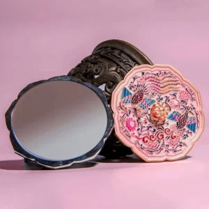 Twin phoenix Chinese pocket mirror opened at an angle on a pink background, showing the colourful floral fenghuang lid, rose gold central ornament, folding mirror, and vintage-inspired decorative styling