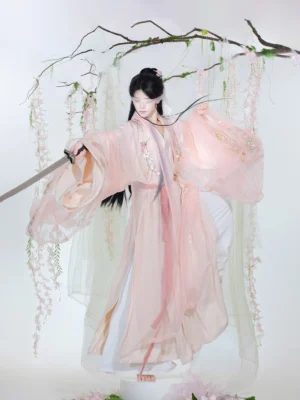 Three-quarter front view of a soft pink embroidered unisex Xianxia hanfu set with wide flowing sleeves, matching waist sash, traditional sword styling, and floral garden backdrop