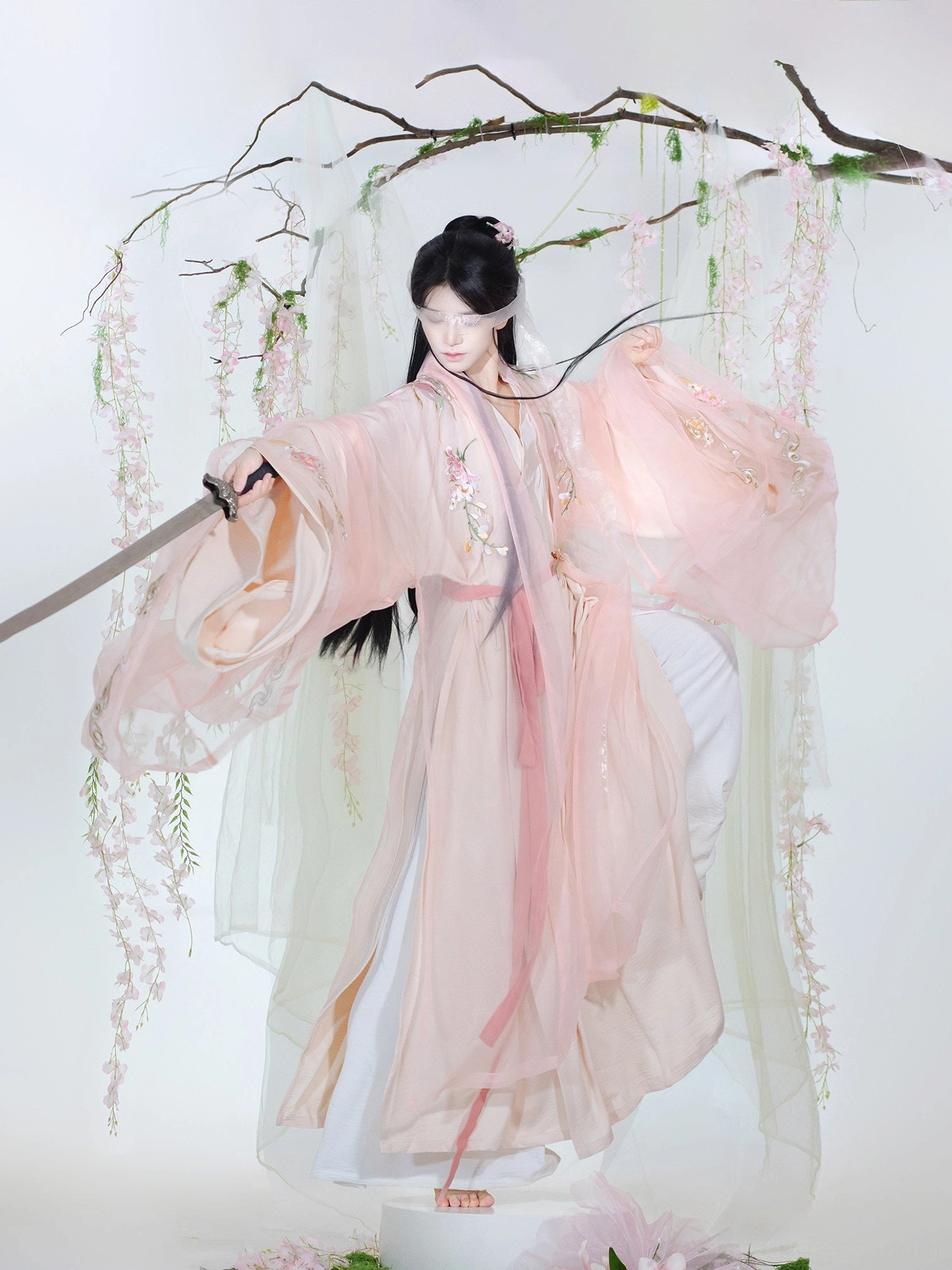 Three-quarter front view of a soft pink embroidered unisex Xianxia hanfu set with wide flowing sleeves, matching waist sash, traditional sword styling, and floral garden backdrop