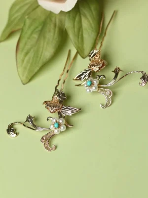 Two vintage Chinese floral hair pins with gold-toned carved flower and leaf designs, imitation pearls, and turquoise-colored beads displayed on a light green background with foliage