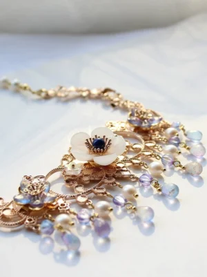 Traditional Chinese Hanfu necklace with rose-gold metal frame, white shell flower centerpiece, dark blue flower center, and blue-purple gradient bead tassels displayed on light fabric