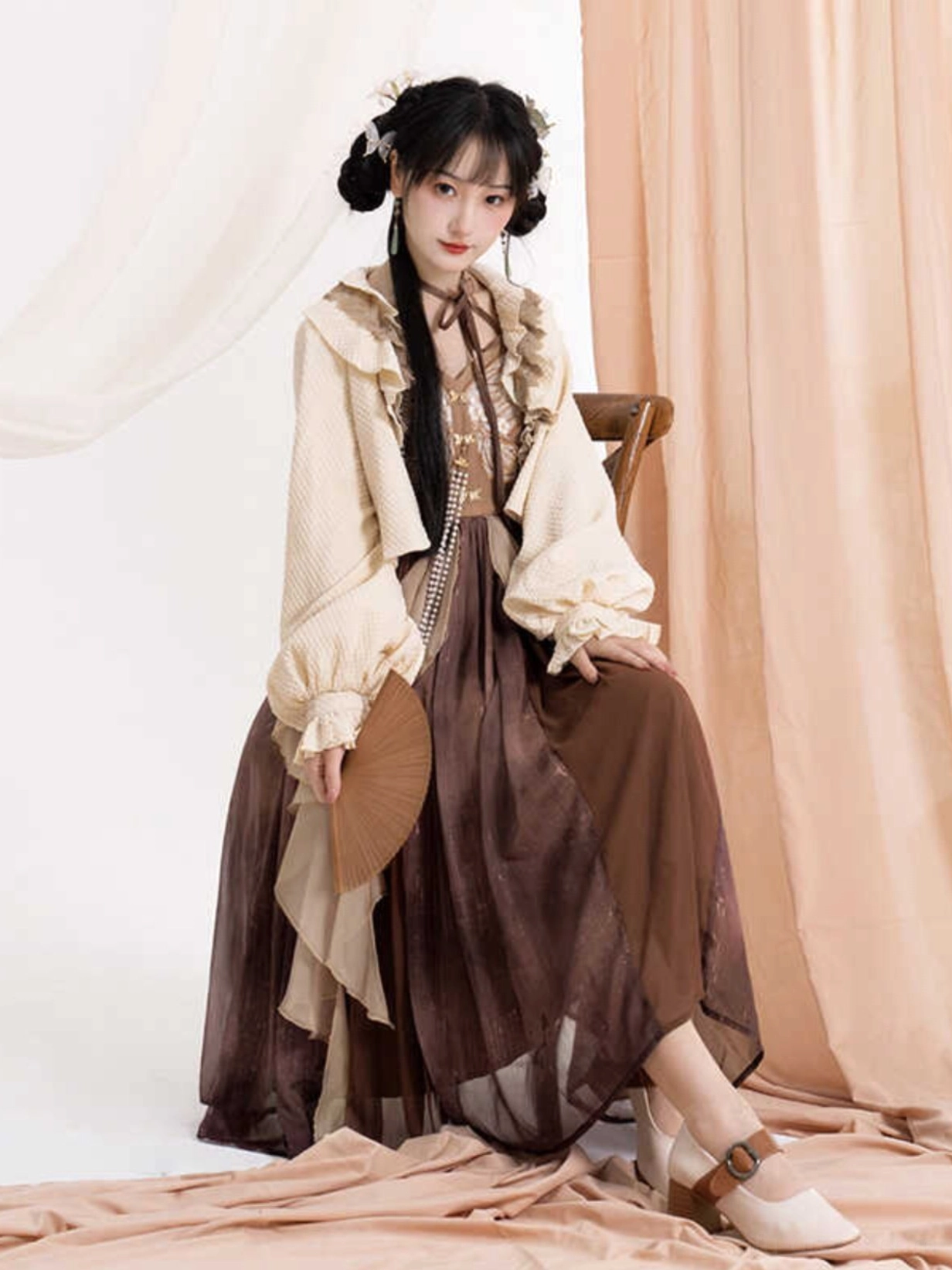 Seated model wearing a vintage Chinese style Hanfu slip dress and cardigan set with pearl tassel decoration, ruffled layers, and folding fan