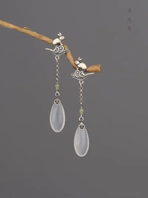 Vintage Chinese rabbit jade drop earrings hanging from a twig, showing aged silver rabbit and cloud studs, green jade beads, delicate chains, and translucent white teardrop pendants