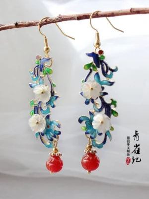 Vintage Chinese style dangle earrings featuring cloisonne floral frames, white shell blossoms, red carved bead pendants, and elegant gold-tone hooks hanging from a natural branch