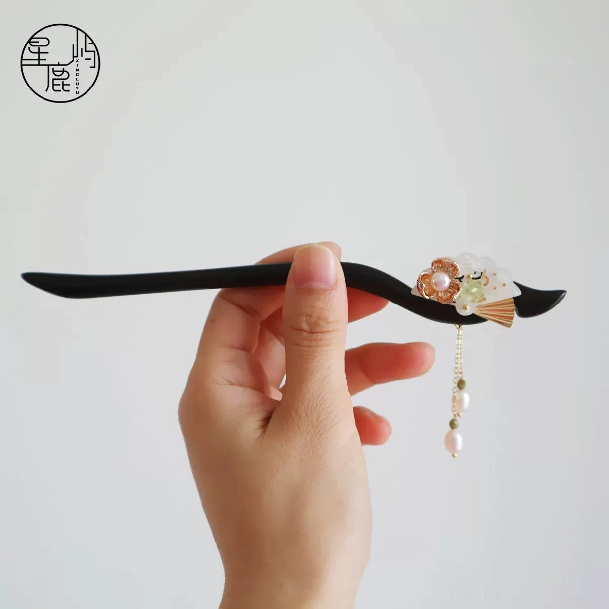 Hand-held view of a vintage Chinese wooden hair stick featuring a polished black shaft, floral decoration, green bead accents, and matching pearl tassel