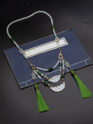 Full-length view of a green and white beaded Chinese necklace with layered chains, decorative pendants, and tassels arranged on a vintage book and dark background